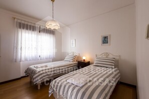 Ein Schlafzimmer mit zwei Einzelbetten, einem Nachttisch mit Lampe, einem Fenster mit durchscheinenden Vorhängen und einem Bild an der Wand.