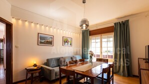 A dining area with a wooden table and chairs, a sofa, a small side table with a lamp, and a window with curtains.