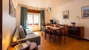 A spacious living area with a sofa, dining table, and chairs. There is a TV, a lamp, and a plant on the table.