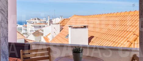 A balcony with two wooden chairs and a small table, overlooking a cityscape with terracotta rooftops.