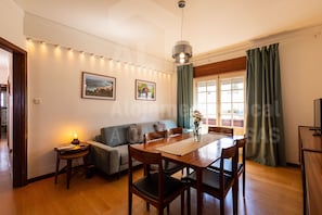 Un coin repas avec une table et des chaises en bois, un canapé, une petite table d’appoint avec une lampe et une fenêtre avec des rideaux.
