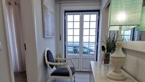 A spacious hallway with a wooden floor, a white chair, a desk with a lamp, and a large window with a view of parked cars. - Ribas Apartment by Acasadascasas (Mafra)