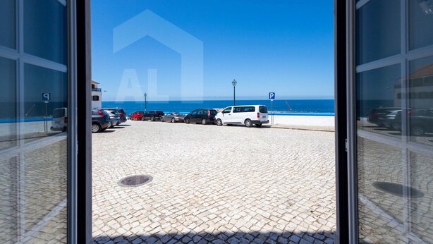 A view through a glass door of a parking area with a cobblestone floor, leading to a beach with parked cars and a clear blue sky. - Ribas Apartment by Acasadascasas (Mafra)