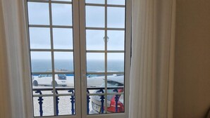 A hotel room with a bed, two rolled towels, and a view of the sea and parked cars through a window. - Ribas Apartment by Acasadascasas (Mafra)
