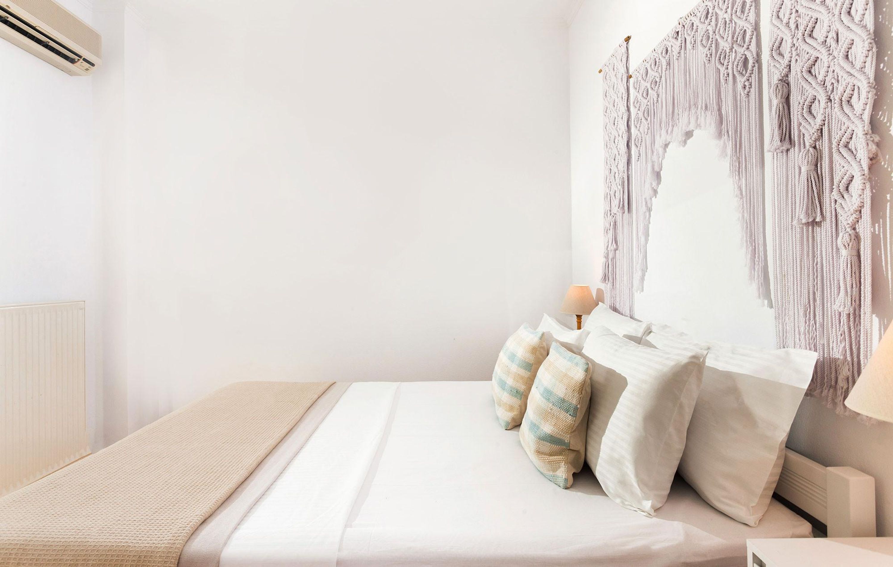 A neatly made bed with a beige blanket and pillows, a macrame wall hanging, and a bedside lamp.