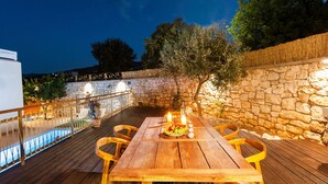 Terraza de madera al aire libre con mesa y sillas, rodeada de muros de piedra y una piscina.