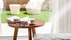 Una mesa redonda de madera con un libro, gafas y una taza, cerca de una ventana que da a un espacio verde con sillas blancas.