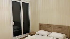 Une chambre à coucher avec un grand lit, une tête de lit en bois, un luminaire blanc au plafond et une porte-fenêtre coulissante.