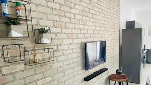 A modern living room with a brick wall, a flat-screen TV mounted above a wooden coffee table, and a minimalist shelving unit with decorative items.