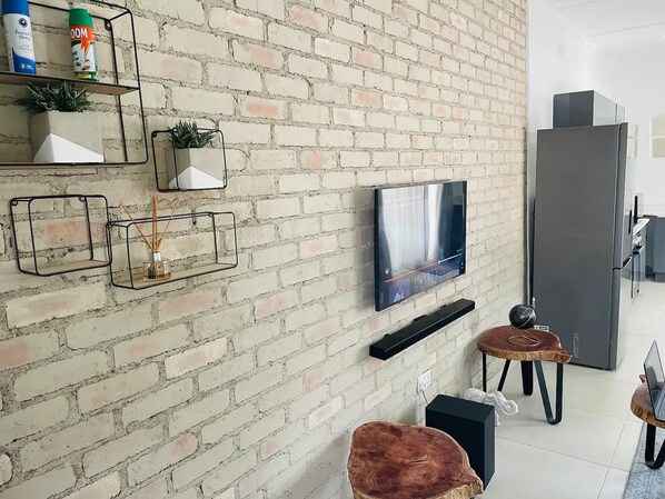 Un salon moderne avec un mur en briques, un téléviseur à écran plat fixé au-dessus d’une table basse en bois, et une étagère minimaliste agrémentée d’objets décoratifs.