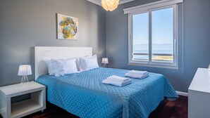 Un dormitorio con una cama grande, una mesita de noche, un escritorio, una ventana con vistas a la playa y una lámpara de techo.