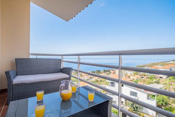A balcony with a wicker sofa, a glass table with drinks, and a scenic view of the sea and buildings.