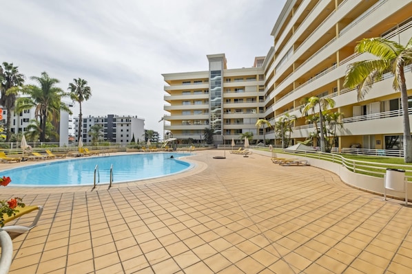 A swimming pool area with tiled flooring, sun loungers, and palm trees.