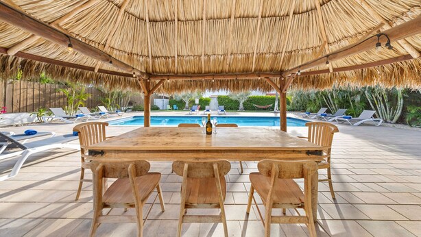 Un área de comedor al aire libre con techo de paja, piscina y sillones de descanso.