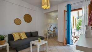 A modern living room with a grey sofa, a white coffee table, and a dining area with a small table and chairs. There is a kitchen visible in the background, and a balcony with blue curtains. - Apartments for 4 1st Floor (Malevizi)