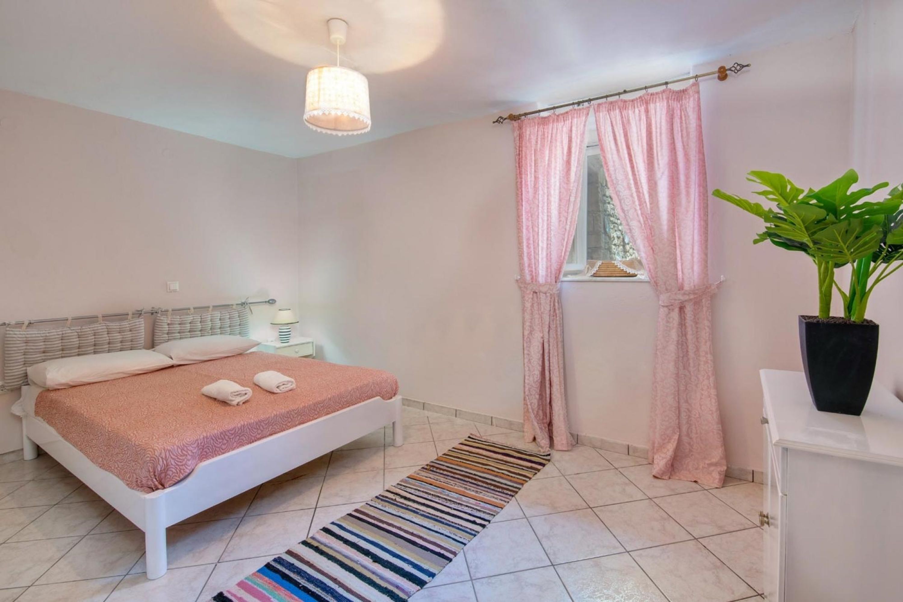 A bedroom with a bed, a bedside table, a lamp, a window with curtains, a potted plant, and a striped rug.