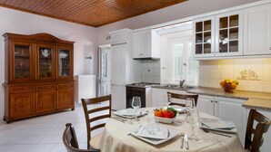 A dining area with a table set for two, a wooden cabinet, and a kitchen with white cabinets and a fruit bowl on the counter. - Angelina Villa - Four Bedroom Traditional Paxos Villa (Paxos)