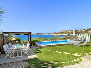 A pool area with a view of the sea, a covered seating area, and a table with chairs.