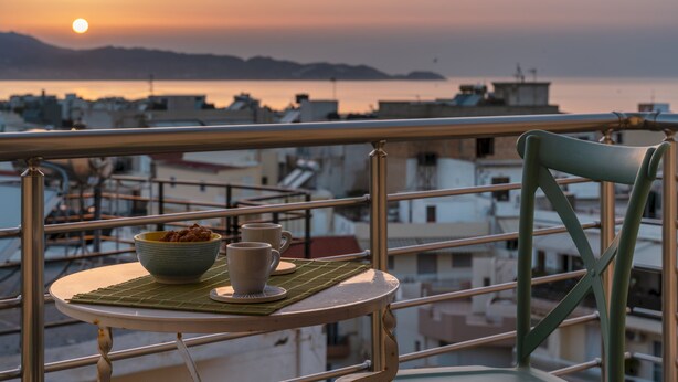 A balcony with a table, a bowl, and two cups, overlooking a cityscape and water during sunset.