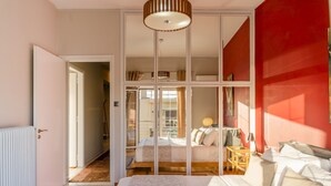 A bedroom with a large bed, a wooden floor, a red accent wall, and a view into a connected room.