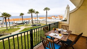 A balcony with a table set for breakfast, overlooking a coastal area with palm trees and buildings. - Kiko s House by Acasadascasas (Mafra)