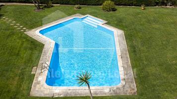 An aerial view of a swimming pool with a geometric shape, surrounded by a well-maintained lawn and a hedge.
