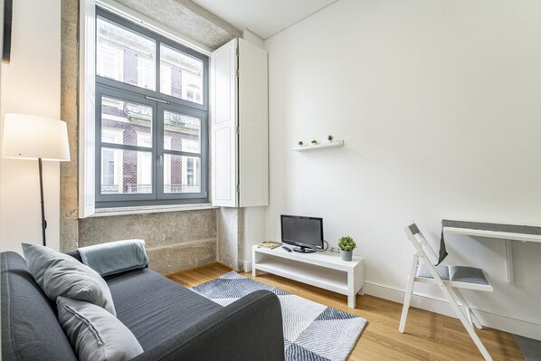 Uma sala moderna com um sofá cinzento, um pequeno móvel branco para TV, uma escrivaninha com uma cadeira e uma janela com vista para edifícios.