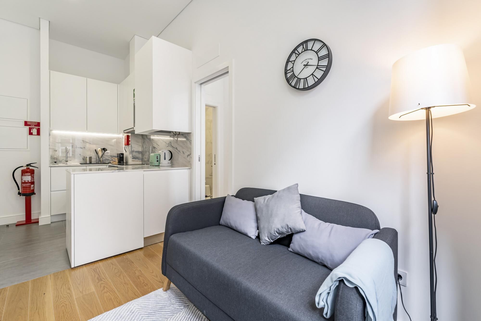 A modern living room with a grey sofa, a wall clock, and a floor lamp.