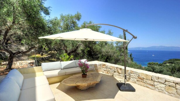 A patio with a white sofa, a round wooden table with a flower, and a large umbrella. - Apeiron II Villa - Sunny Modern Pool - Walk to Gaios (Paxos)