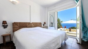 A bedroom with a large bed, a nightstand, a lamp, and a view of the sea through an open door. - Apeiron II Villa - Sunny Modern Pool - Walk to Gaios (Paxos)