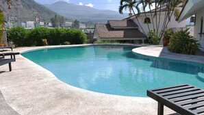 A swimming pool with a curved edge, surrounded by a concrete deck. There are lounge chairs and potted plants nearby. In the background, there is a building with a tiled roof and a mountainous landscape. - Hotel Hacienda las Higueras  (Tepic)