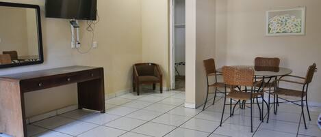 Una habitación con un escritorio, un televisor montado en la pared, una mesa de comedor con sillas y una puerta que da a otra habitación.