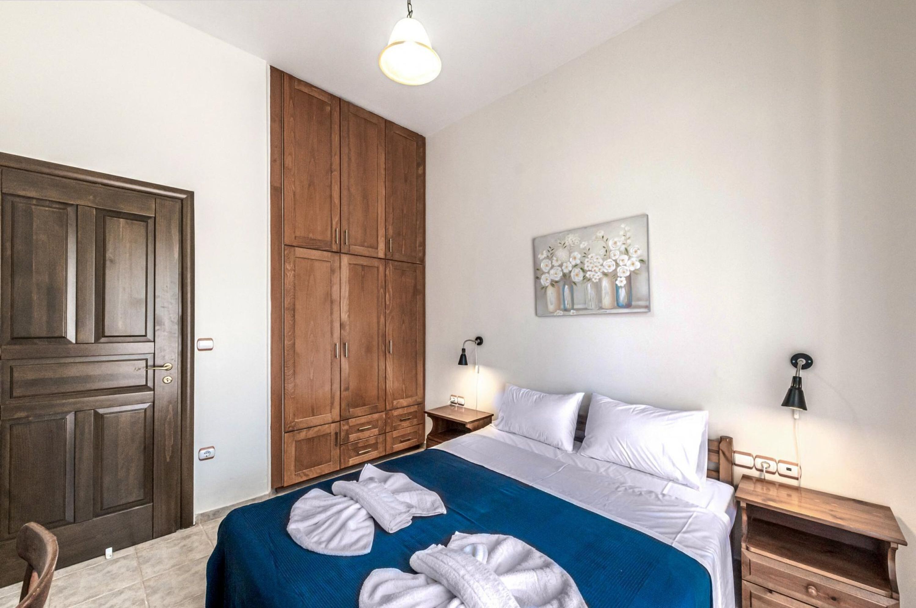Habitación de hotel con un amplio armario de madera, cama con sábanas blancas y una colcha azul, además de un cuadro en la pared.