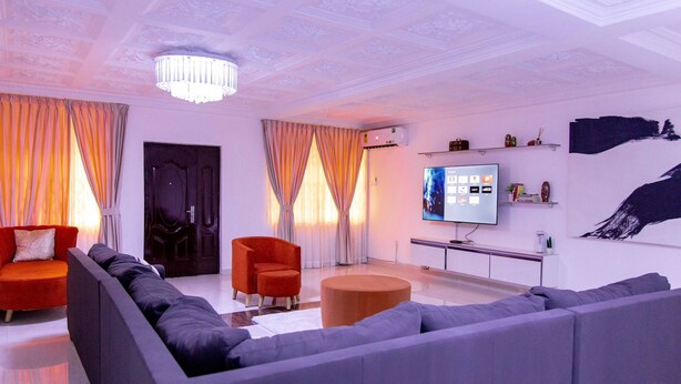 A modern living room with a grey sofa, red armchair, and orange ottoman. A flat-screen TV is mounted on the wall, and there are decorative items on shelves. - Family sized house with a summer hut for outdoor sitting (Tema)