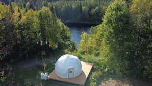 A geodesic dome structure with a wooden deck, surrounded by dense forest and a river. - Dome # 1 with view to the mountains (Saint-Gabriel-de-Valcartier)