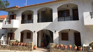 A white building with multiple balconies, stone walls, and terracotta pots. - Nikos Apartments A2 in Gialiskari (Ikaria)