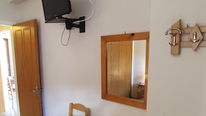 A room with a wooden chair, a small wooden table with a folded white towel, a wall-mounted TV, and a wooden wardrobe. - Nikos Apartments A2 in Gialiskari (Ikaria)
