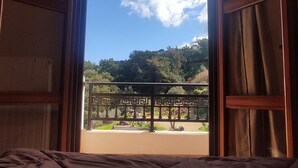 A room with a bed, an open window, and a view of a garden and trees. - Nikos Apartments A2 in Gialiskari (Ikaria)