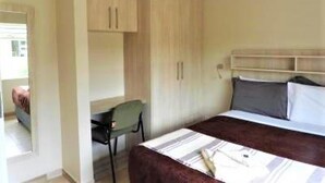 A bedroom with a bed, a desk, and a chair. There is a wardrobe with two doors and a shelf above the bed.