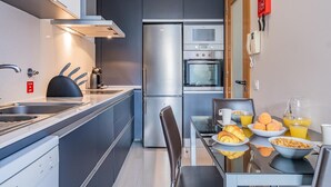 A compact kitchen with a refrigerator, oven, and microwave. There is a dining table with breakfast items and a chair. - Living Funchal by Homie (Funchal)