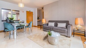 A modern living room with a grey sofa, two side tables with lamps, a dining table with chairs, and a large window with curtains. - Living Funchal by Homie (Funchal)