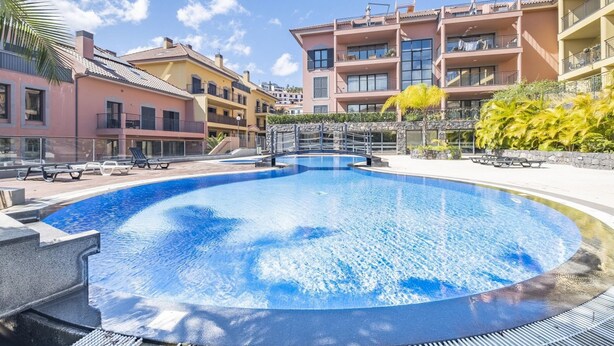 A swimming pool surrounded by multi-story buildings with balconies, a stone wall, and palm trees. - Living Funchal by Homie (Funchal)