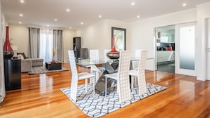 Un salon moderne avec une table à manger en verre, des chaises blanches, un canapé et une cuisine en arrière-plan.