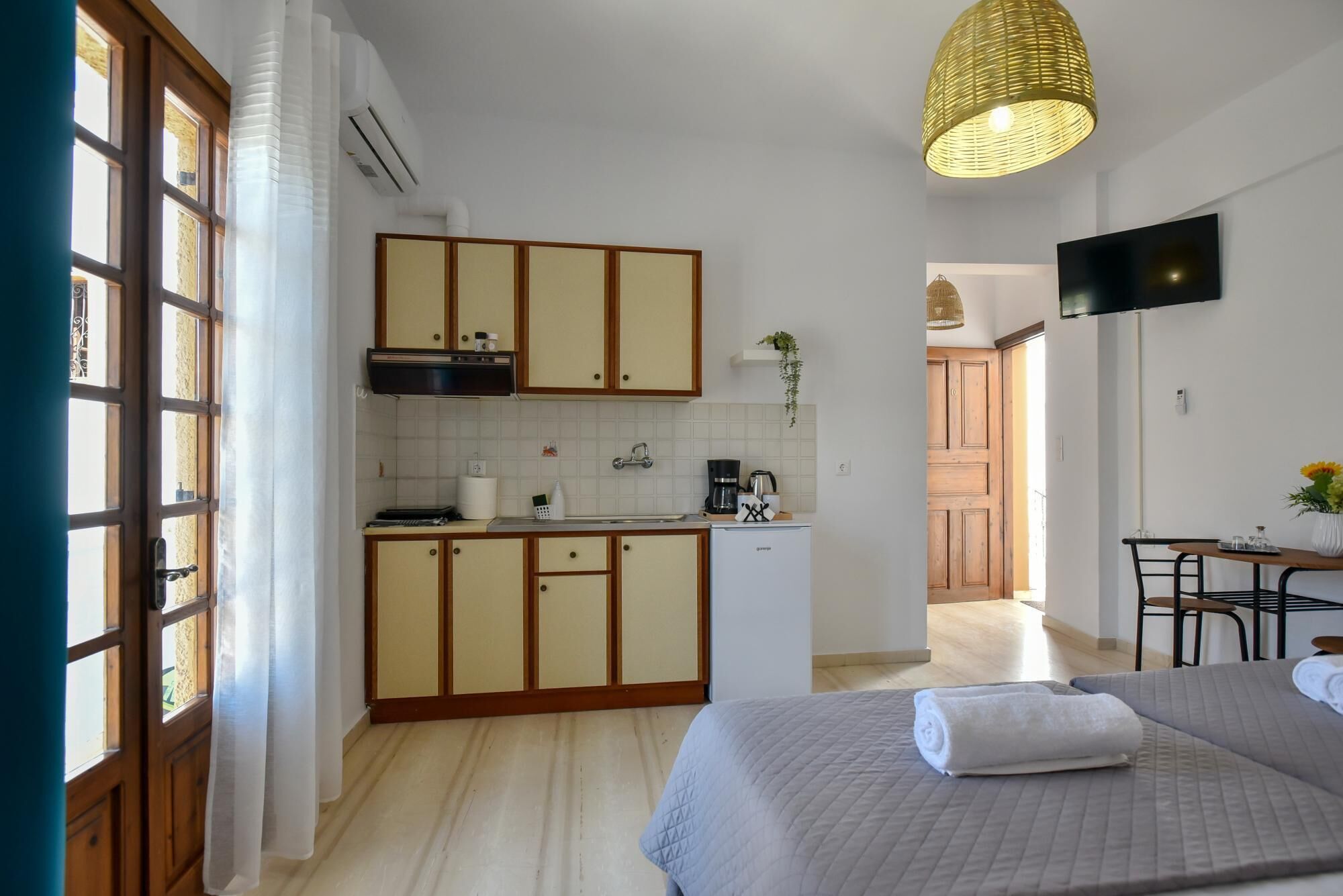 A compact hotel room with a kitchenette, a bed with a rolled-up towel, a television, and a hanging light fixture.
