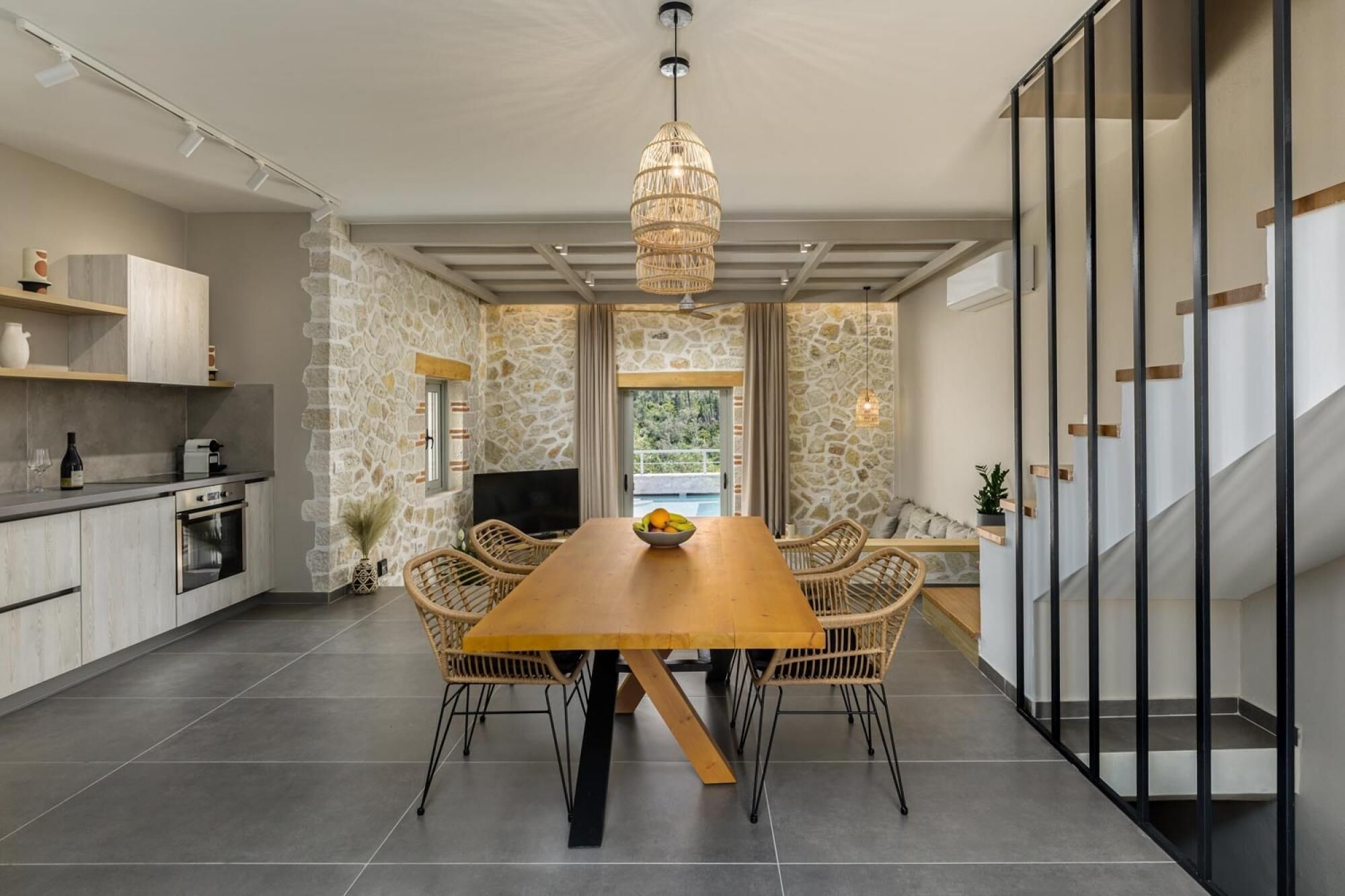 A modern kitchen with stone walls, a dining area with a wooden table and wicker chairs, and a staircase with a glass railing.