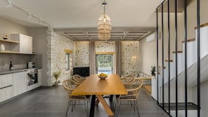 Une cuisine moderne aux murs en pierre, un coin repas avec une table en bois et des chaises en osier, et un escalier doté d’une rampe en verre.