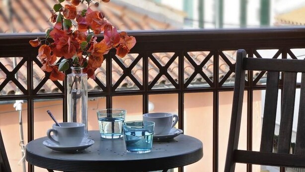 Un balcón con una mesa redonda, dos sillas, un florero con flores y vistas a edificios y árboles.