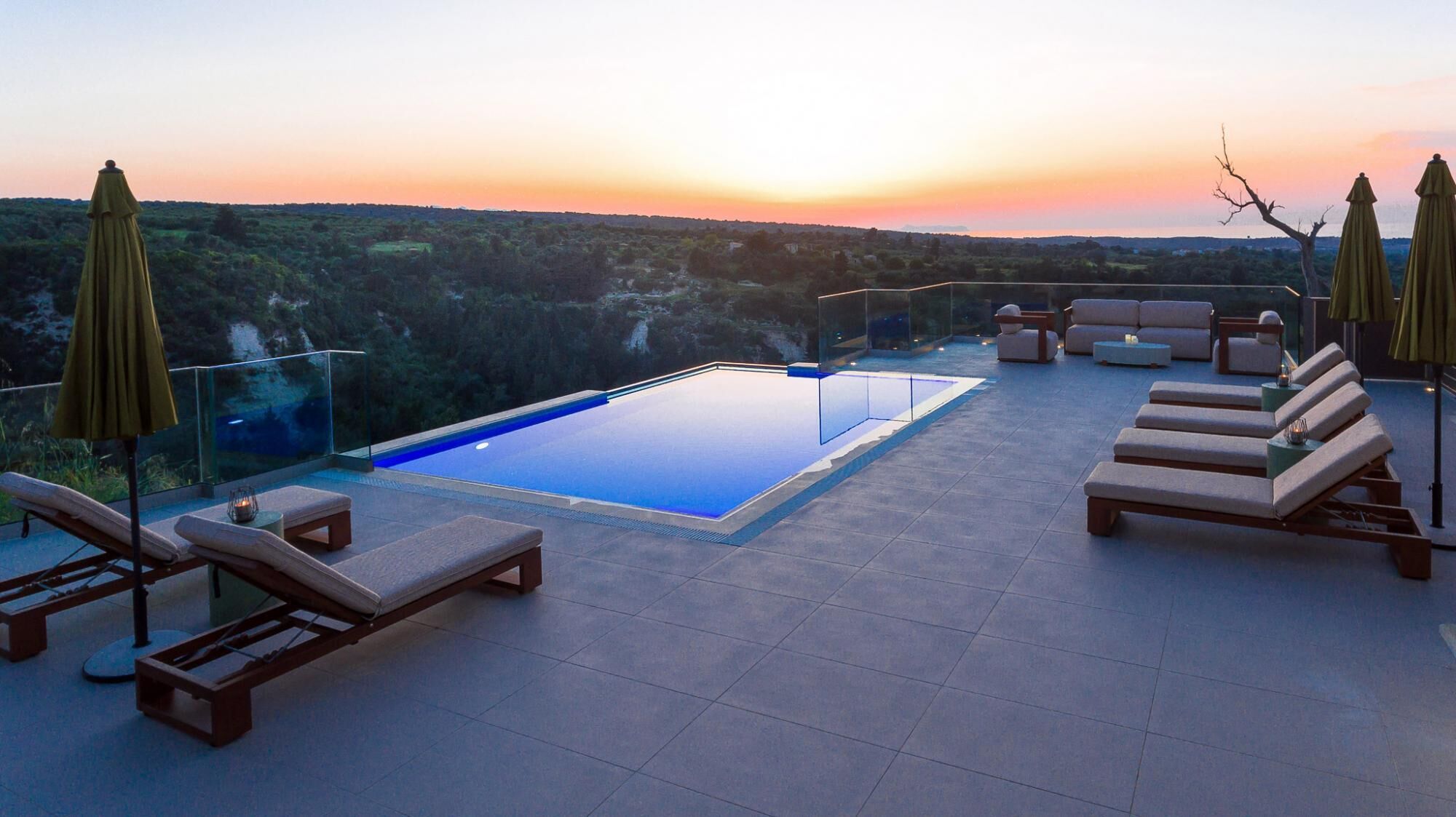 A rooftop infinity pool with lounge chairs and umbrellas, surrounded by a scenic view of hills and a sunset.