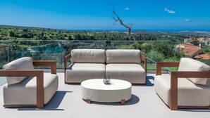 A terrace with a sofa set and chairs, a round table, and a glass railing overlooking a landscape with trees and buildings. - Dim Luxury Villa - With Private Pool (Mylopotamos)