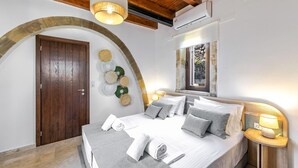 A bedroom with a wooden door, a bed with pillows and a blanket, a bedside table with a lamp, and a mirror on the wall. - Dim Luxury Villa - With Private Pool (Mylopotamos)